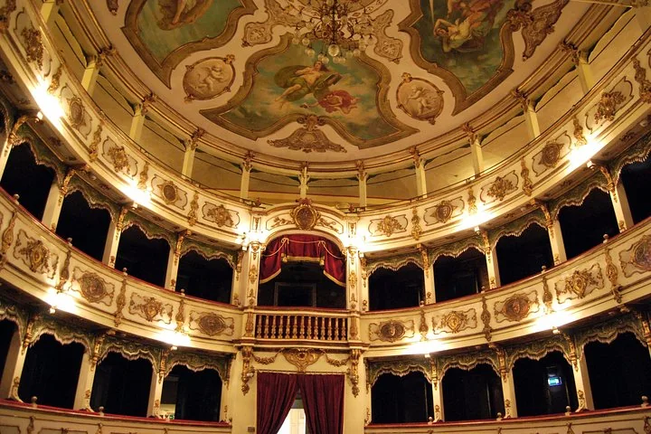 The horseshoe hall of the Teatro Verdi in Busseto with cream and gold tiers of boxes, a draped central box and a painted ceiling