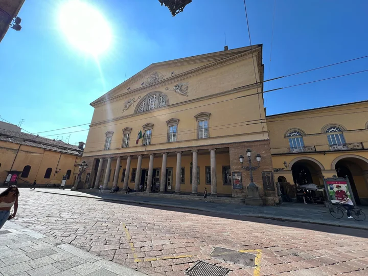 The yellow facade and colonnade of the Teatro Regio on a sunny day, seen across a paved street