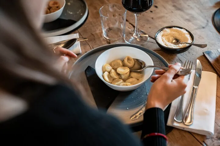 Ein Gast löffelt kleine gefüllte Nudeln aus einer weißen Schale, mit geriebenem Parmigiano und einem Glas Rotwein auf einem Holztisch