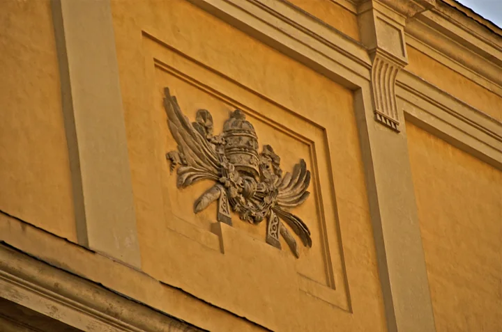 A carved relief of a crown framed by palm fronds and ribbons, in a panel on the yellow facade