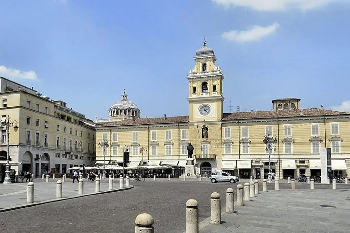 Die Piazza Garibaldi in Parma mit einem gelben Palazzo und seinem Uhrturm, einer Statue und einer Reihe steinerner Poller unter blauem Himmel