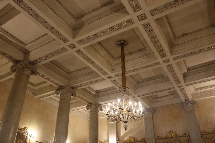A pale foyer with stone columns, a coffered ceiling and a crystal chandelier hanging on a long rod