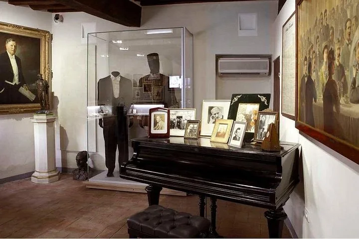 Ein Museumsraum mit einem schwarzen Flügel, gerahmten Fotografien, einem Porträt in vergoldetem Rahmen und einer Vitrine mit einem Anzug