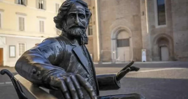Eine Bronzestatue eines bärtigen Verdi, der mit erhobener Hand auf einer Bank sitzt, vor hellen und backsteinernen Gebäuden