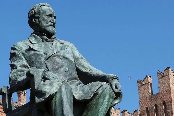 Eine sitzende Bronzestatue Verdis vor blauem Himmel, dahinter die Zinnen einer Backsteinburg